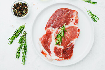 Uncooked pork steak on white plate with rosemary and pepper