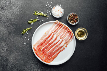 Raw bacon slices on white plate with pepper, salt and rosemary