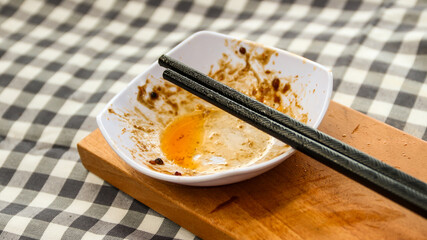 An empty white dish with food remnants and chopsticks resting on top, placed on a wooden board over a checkered tablecloth