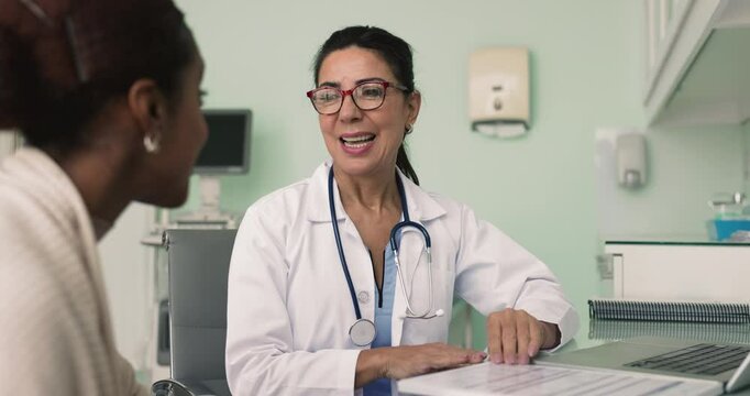 Senior Latin doctor woman listening to young African female patient at workplace table in office, nodding, talking, asking questions, meeting for consultation, examination