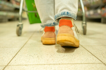 Legs of buyer with shopping cart walking in supermarket close-up rear view.