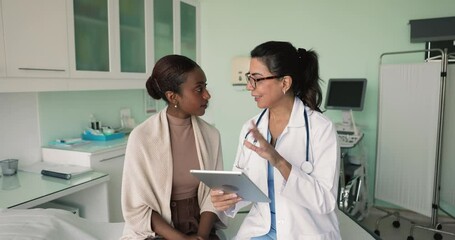 Mature female doctor talking to young African patient after checkup, showing digital tablet, electronic content, explaining diagnosis, treatment, examination result, sitting on couch - Powered by Adobe