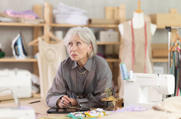 Mature dressmaker draws a sketch of clothes and patterns on an electronic tablet. Woman creates a...