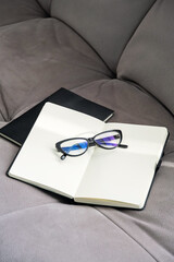 Pair of Stylish Black Glasses Rests on an Open Notebook Placed on Soft Gray Couch.