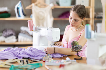Young dressmaker sitting and sewing fabric on a sewing machine. Seamstress in the workshop. Process of tailoring clothes according to individual measurements and sketches