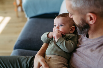 Father holding baby boy in arms, sitting with him on sofa.