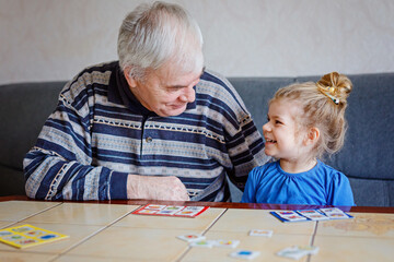 Beautiful toddler girl and grandfather playing together pictures memory table cards game at home. Cute child and senior man having fun together. Happy family indoors
