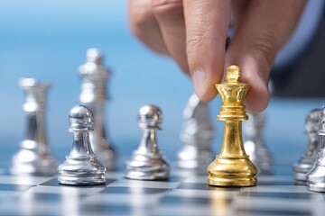 Businessman moving a golden chess piece on a board, with other silver pieces in the background. He is wearing a blue suit 