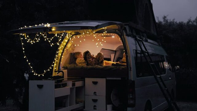 Cozy van interior with fairy lights, two people on a bed, and soft blankets in a peaceful evening atmosphere.