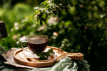 A serene outdoor setting with a glass cup of black coffee on a wooden tray, adorned with a small...