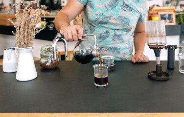 Unrecognizable barista expertly pours brewed coffee from Japanese siphon coffee maker into a glass, showcasing the art of specialty coffee preparation. Copy space at the bottom of image.