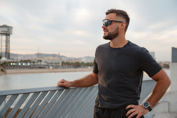Sportsman wearing sunglasses, relaxing after training, gazing at the beautiful Barceloneta beach and the shimmering sea in Barcelona