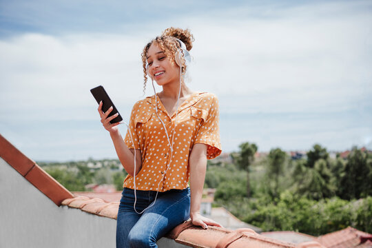 Smiling woman using smart phone while sitting on railing in front of sky