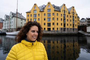Portrait of a young brunette woman looking away, wearing a yellow anorak, with the Brosundet Canal and singular building in Alesund, Norway, behind her