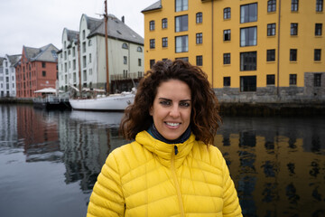Portrait of a young brunette woman looking at the camera, wrapped in a yellow down jacket, with the Brosundet Canal in Alesund, Norway, behind her, with art nouveau buildings