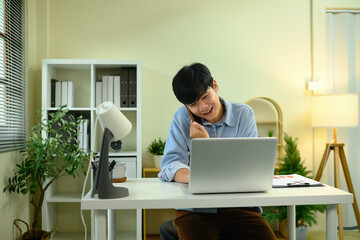 Happy young entrepreneur talking on phone while working on laptop at desk
