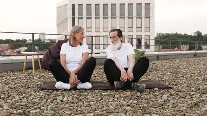 Senior man and woman seated on rooftop performing yoga exercises outdoors. Represents meditation, health, relaxation, and urban wellness. Models are older adults in casual attire