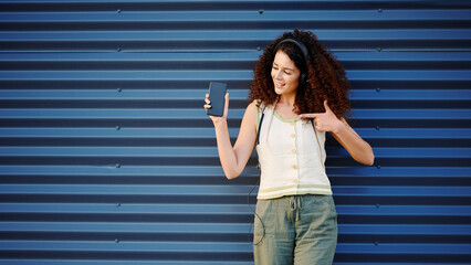 woman with smartphone smiling joyfully against blue wall backdrop, wearing headphones, enjoys music...