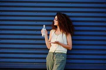 woman holding water bottle leans against blue wall. casual fashion, curly hair, and colorful...