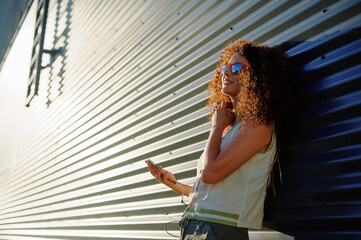 woman with smartphone and sunglasses leans casually against industrial corrugated wall, sunlit hair...