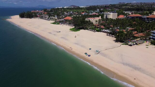 Drone flies right to left in a half-circle keeping the beach centered, revealing luxury resorts on the shore and the expansive horizon, capturing the stunning coastal landscape.