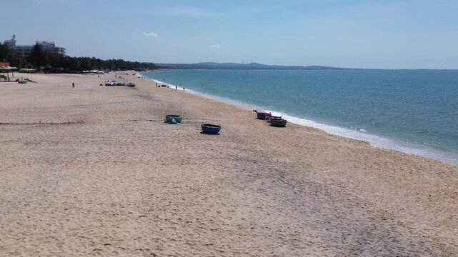Drone flies in a half-circle from right to left, keeping Mui Ne beach centered with fishing buckets on sand, revealing resorts, white shore, and the sea horizon.