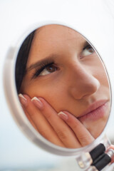 A young woman holds a round mirror, looking at her reflection. She is touching her cheek and posing for social media, showcasing a fashionable and modern aesthetic