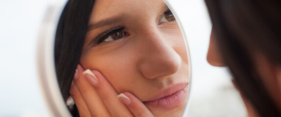 A young woman looks at her reflection in a mirror, gently touching her face with her hand. She is inside. The natural light suggests it is daytime. Her nails are manicured