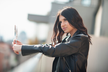 A fashionable young woman in a black leather jacket leans against a balcony railing and gazes into a hand mirror. She is on the balcony of an apartment, posing for social media
