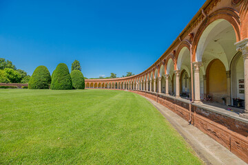 Ancient cemetery cloister featuring hanging lamps, tombs, and sculptures in the 15th-century Charterhouse of Ferrara, set against a lush green lawn, in Ferrara city of Italy