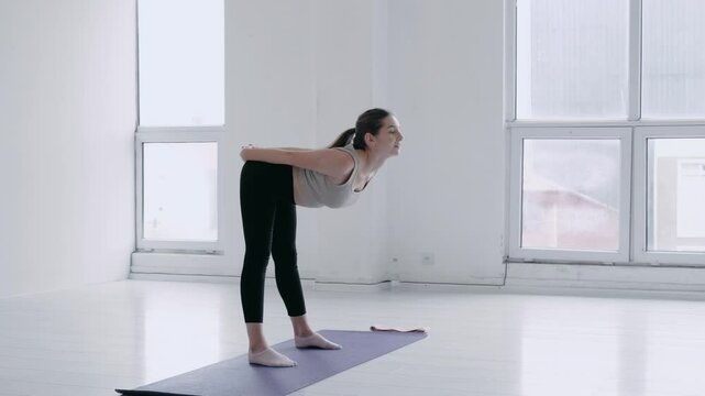 Healthy wellness caucasian girl with long hair in sportswear doing warm-up before main exercise or yoga In a bright studio with windows. Woman arching and stretching the muscles of the back and neck.
