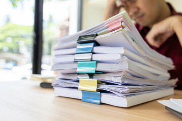 A man in a red shirt is sitting at a desk with a pile of papers in front of him. He is stressed and...