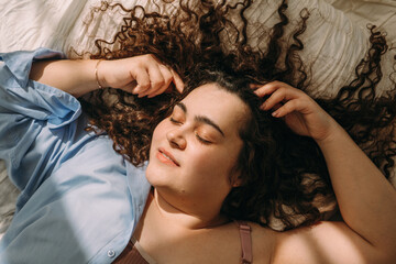 Fototapeta premium A portrait of a young, attractive plus-size woman with dark curly hair lying in bed with her eyes closed.