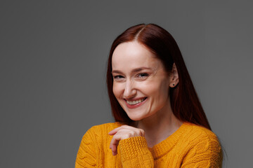 Happy red haired woman in sweater looking at camera isolated on grey