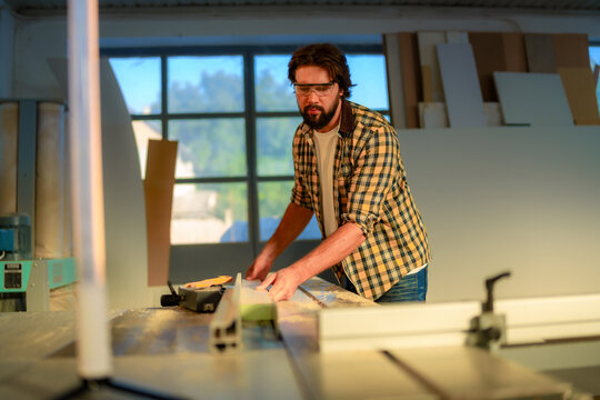 Professional carpenter wearing safety glasses carefully cutting wooden plank using circular saw in well lit workshop, demonstrating precision and expertise in woodworking