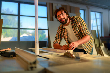 Skilled carpenter wearing safety glasses is carefully measuring a wooden plank in his workshop, preparing for precise cutting using a table saw in the background