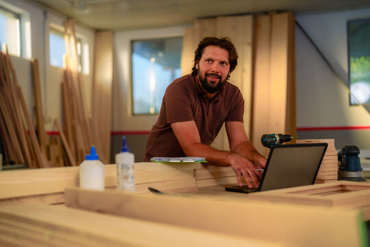 Carpenter working on a laptop in a bustling workshop, surrounded by wood planks, tools, and glue bottles, efficiently managing business tasks and fulfilling orders