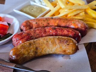 A close-up of grilled sausages served with crispy French fries, mustard dipping sauce, and pickled vegetables. A delicious and hearty meal, perfect for comfort food lovers.