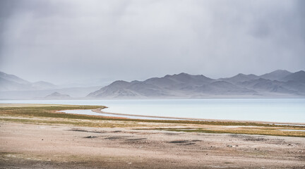Grass and stone shore of Lake Karakul with blue water and mountain peaks in the background, cloudy weather and sky with clouds and clouds in the Pamirs, panoramic landscape