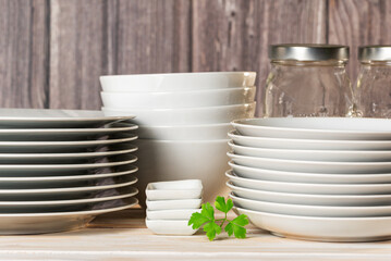 White tableware with plates and bowls next to some glass jars, on a light wooden background, close-up.
