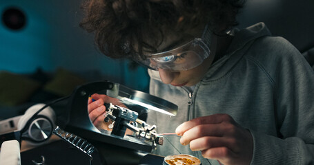 A school-age boy wears safety glasses while repairing a computer motherboard. He solders cables and works with microprocessors, learning modern electronics with focus and precision.