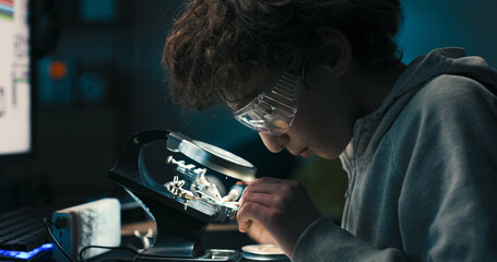 Focused on the task, the child solders tiny wires on a circuit board. Wearing goggles, he explores the basics of electronics, repairing components with care and precision.