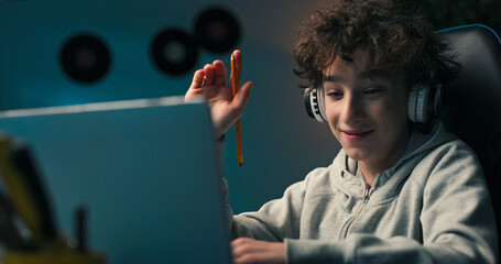 A boy with curly hair sits at a desk wearing wireless headphones, taking notes and redrawing. He...