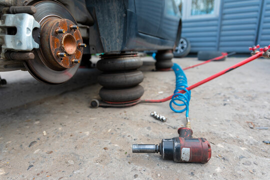 Car lifted with air jacks, brake hub exposed, with pneumatic impact wrench and lug nuts on the ground in a repair setting. - Powered by Adobe