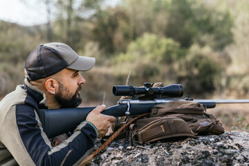 Hunter aiming with rifle lying on a rock in nature