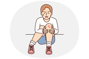 Crying boy sits on ground and holds on to injury on knee after fall during walk