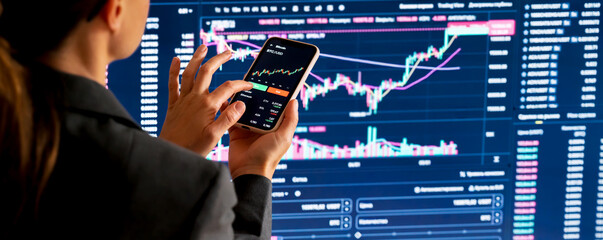 Rear view of businesswoman trading cryptocurrency on smartphone with financial charts in background
