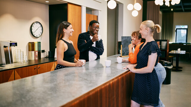Casual Office Meeting with Colleagues Enjoying Coffee Break at Work. They are standing relaxed in a canteen. There is a happy and positive vibe around them.