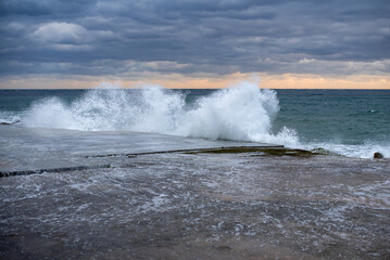 Obraz premium Stormy sea waves crashing against a rocky shore at sunset. Dark clouds fill the sky, creating a dramatic atmosphere with vibrant colors on the horizon.