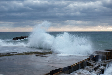 Stormy sea waves crashing against rocks at sunset. Dark clouds fill the sky, creating a dramatic atmosphere. The ocean reflects the fading light.
